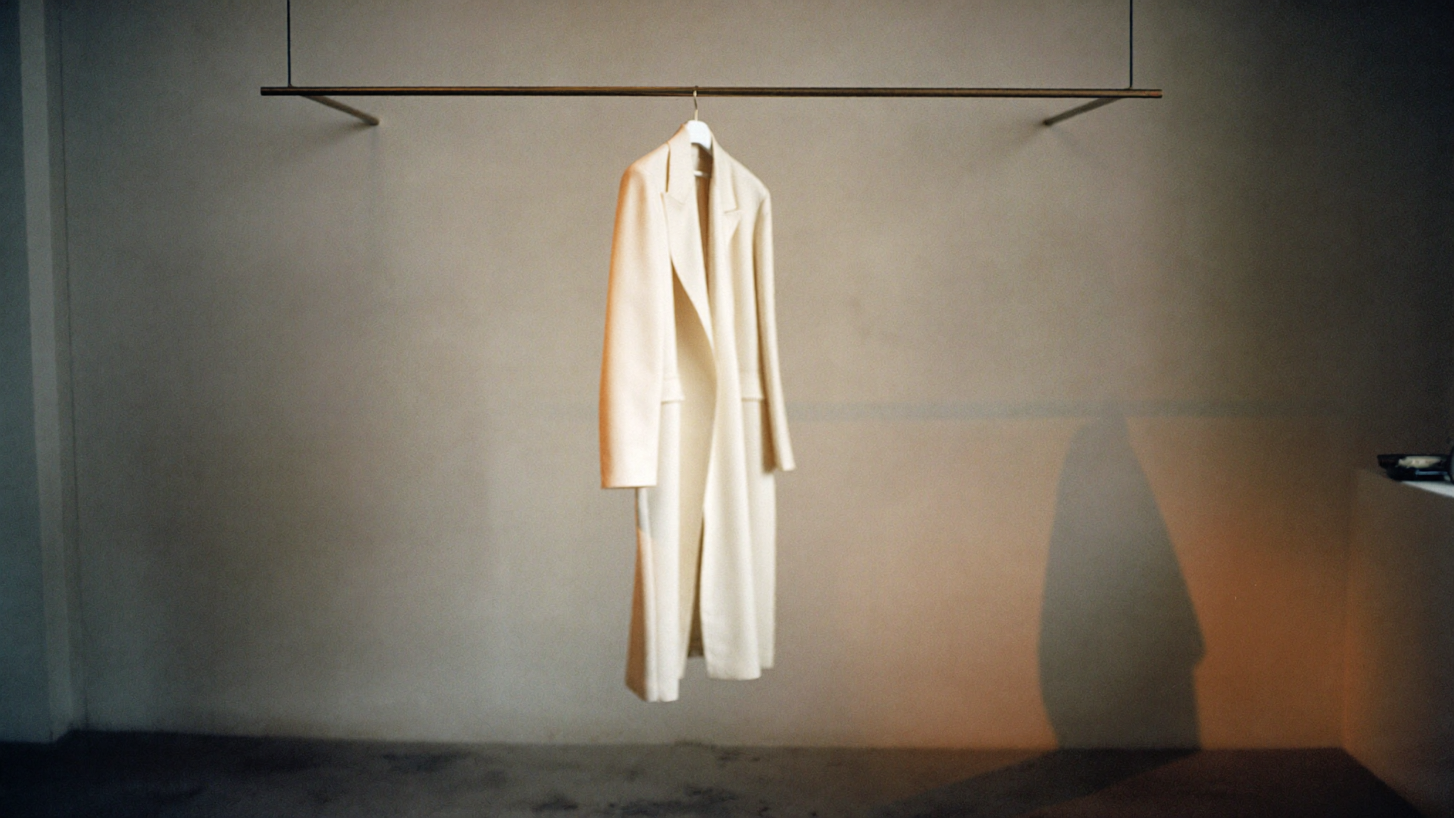 Leeres, minimalistisches Atelier in der Dämmerung mit einem ivory-farbenen Mantel an einer schlanken Messingstange.