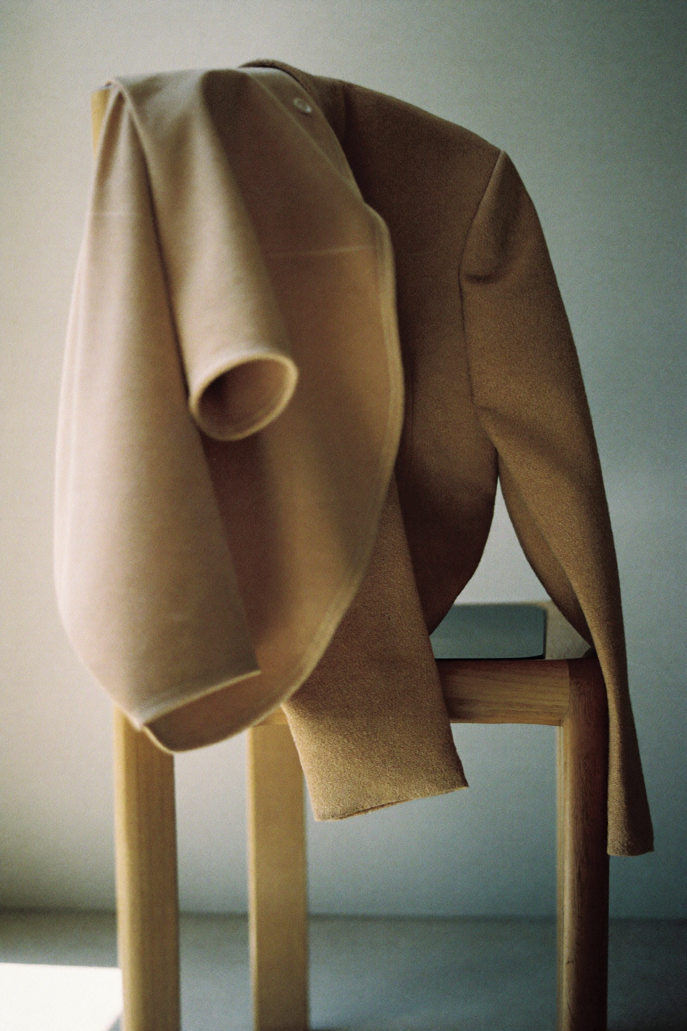 Kamelfarbener Wollblazer über einem minimalistischen Holzstuhl vor einer schlichten Wand.